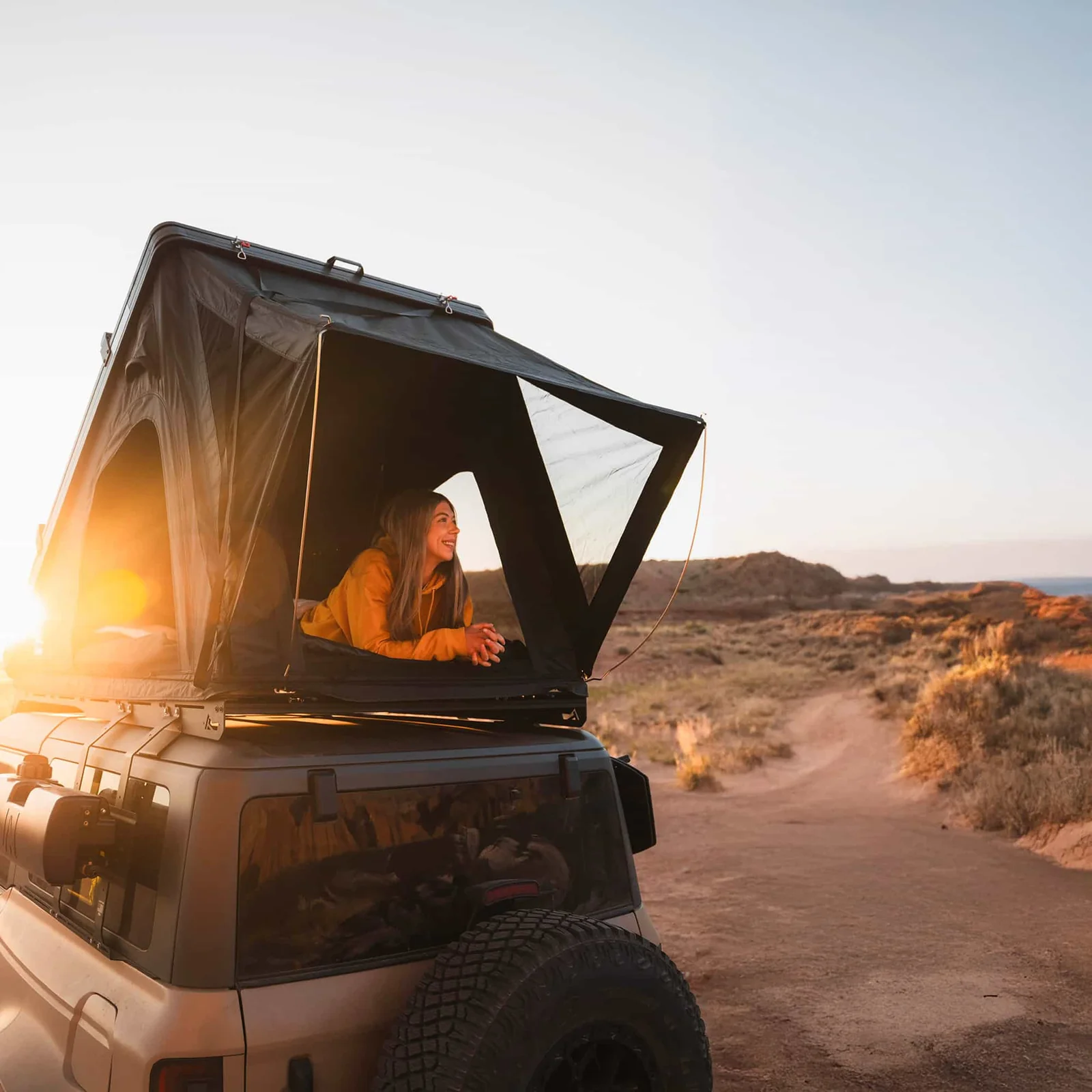 Femme souriante dans une tente de toit rigide sur un 4x4 au coucher de soleil en paysage désertique