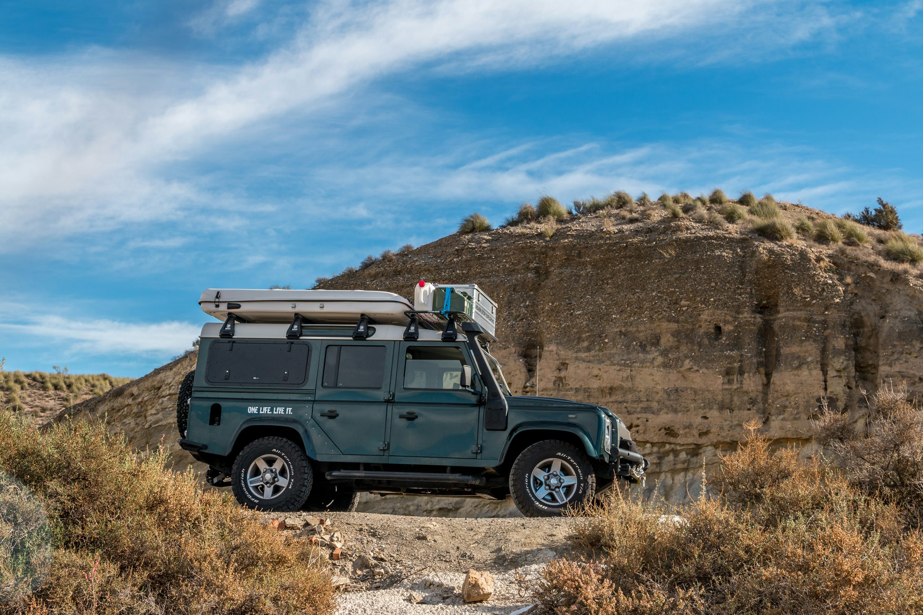Land Rover Defender 4x4 équipé d'une tente de toit rigide en terrain aride, prêt pour l'aventure overland