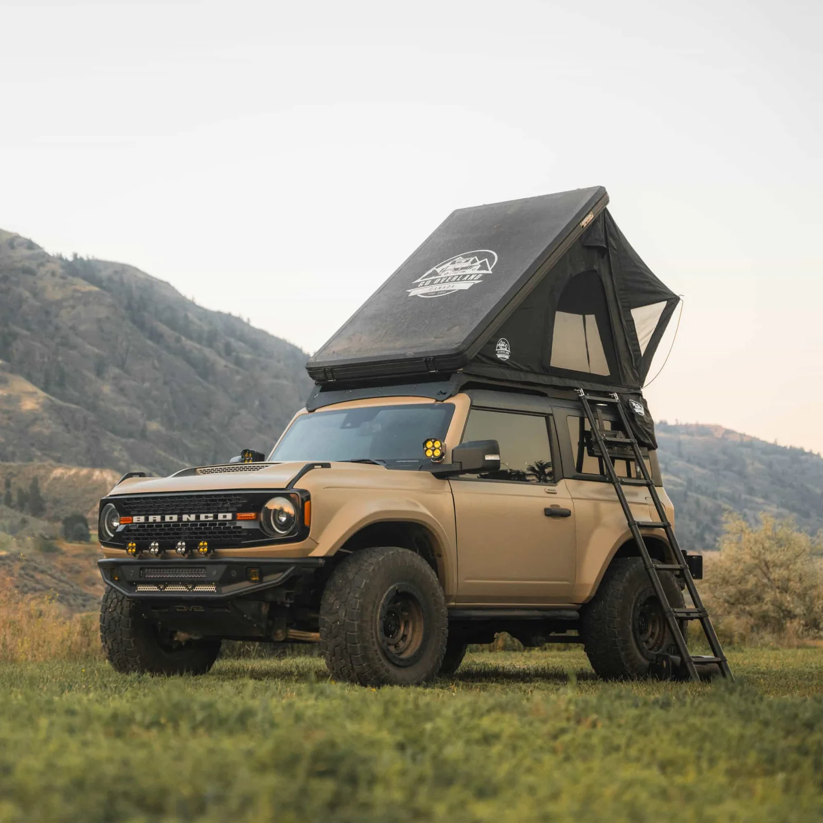 Tente de toit rigide noire déployée sur un Ford Bronco 4x4 beige avec échelle, en pleine nature face aux montagnes